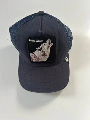 Marinblå keps Goorin Bros Lone Wolf - Snygg marinblå truckerkeps från Goorin Bros med mesh på sidorna och broderad patch framtill med texten 'Lone Wolf' och en ylande varg. Justerbar baktill för perfekt passform. Perfekt accessoar för en cool och avslappnad stil.
