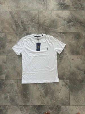 Vit t-shirt från Polo Ralph Lauren - Klassisk vit t-shirt från Polo Ralph Lauren med den ikoniska lilla broderade loggan på bröstet. T-shirten har rund hals, korta ärmar och är tillverkad i mjuk bomull för en skön känsla. Perfekt basplagg till garderoben.