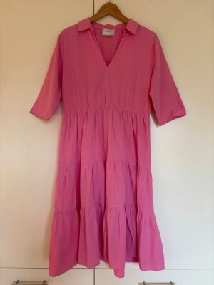 Rosa skjortklänning i midilängd med volanger från Neo Noir - En rosa skjortklänning med v-ringning, krage och trekvartsärmar. Klänningen har flera volangvåder som ger en luftig och avslappnad look. Materialet är bomull. Från Neo Noir. Storlek M, passar även S/36
