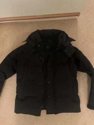 Canada goose jacka  - Säljer en svart pufferjacka med huva och dragkedja samt knappar framtill. Jackan har en patch på ärmen och breda, vadderade paneler som ger en riktigt fet look. Perfekt för kalla dagar när du vill hålla värmen och ändå se stilren ut.
