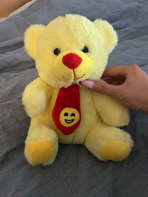 Gul nallebjörn med emoji-slips - Söt gul mjuk nallebjörn med röd nos och svartvita ögon. Nallen har en röd slips med en glad emoji på magen. Perfekt som present eller dekoration i rummet.