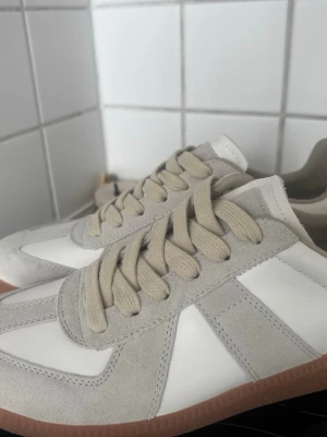 Nya Maison margiela size 42  - Snygga sneakers i vitt och beige med beigea mockadetaljer och matchande snören. Skorna har en klassisk siluett med vit ovandel i läder och beige sula i gummi. Perfekta för en clean och stilren look.