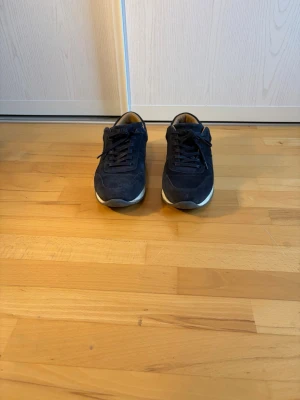 Mörkblå  sneakers i mocka - Säljer ett par mörkblå sneakers i mocka med klassisk låg profil och vita sulor. Skorna har snörning och en stilren design som passar till det mesta. Insidan är ljusbrun och sulan är något upphöjd för extra komfort.
