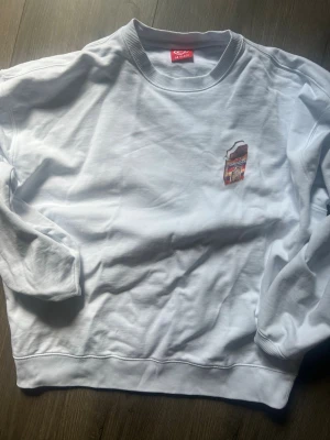 Vit sweatshirt från GB Glace - Säljer en vit sweatshirt från GB Glace med ett litet glassmotiv broderat på bröstet. Tröjan har rund halsringning och ribbade muddar vid ärmslut och nederkant. Perfekt för dig som gillar glass och vill ha en unik tröja i garderoben.