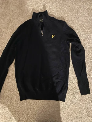 Svart stickad tröja Lyle & Scott - Snygg svart stickad tröja från Lyle & Scott med halv dragkedja och gul logga på bröstet. Tröjan har lång ärm, ribbade muddar och klassisk passform. Perfekt till jeans eller chinos för en clean look.