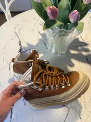 Bruna mockakängor med vita detaljer - Snygga kängor i brun mocka med vita detaljer och beige sula. Skorna har grova snören och metallhakar för en cool outdoor-look. Insidan är fodrad med mjukt material för extra komfort. Perfekta för dig som vill ha både stil och funktion.