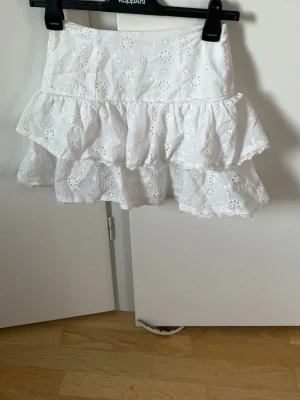 Vit volangkjol med broderie anglaise - Supersöt vit kjol med två volanger och broderie anglaise-mönster. Kjolen är kort och har en feminin, luftig känsla med blommiga broderier. Perfekt till sommaren och lätt att matcha med olika toppar.