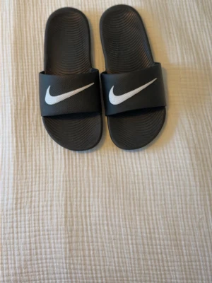 Svarta Nike badtofflor - Säljer ett par svarta Nike badtofflor med vit logga på remmen. Klassisk slip-on modell med räfflad sula för extra grepp. Tillverkade i syntetmaterial som torkar snabbt och är lätta att hålla rena. Perfekta för stranden eller gymmet. Aldrig använda 