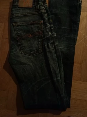 Nudie Mörkblå jeans med bakficka  - Säljer ett par mörkblå jeans med klassisk femficksdesign och snygga slitningar. Jeansen har en bakficka med dekorativ söm och raka ben. Perfekta för en avslappnad och trendig look.