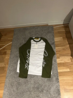 Vit och grön långärmad t-shirt - Snygg långärmad t-shirt med vita och olivgröna partier. Tröjan har raglanärmar i grönt och vitt bröstparti med diskret broderad text på vänster sida. Perfekt för en avslappnad streetwear-look.