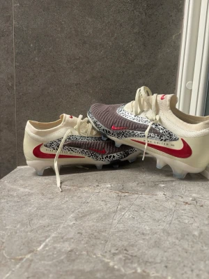Nike fotbollsskor beige/röd/grå - Snygga fotbollsskor från Nike i beige med röda detaljer och gråmelerad ovandel. Skorna har snörning, låg profil och mönstrad sula för bra grepp på planen. Unik design med djurmönstrade inslag och textdetaljer på sidan. Perfekta för dig som vill sticka ut på planen.