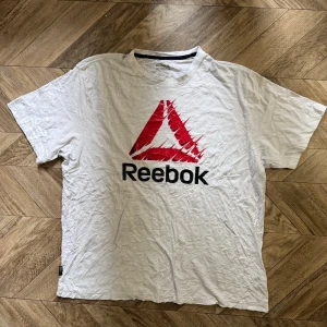 Vit Reebok t-shirt med logga - Vit t-shirt från Reebok med stor röd och svart logga på bröstet. Klassisk passform med korta ärmar och rund halsringning. Tillverkad i mjuk bomull som känns skön mot huden. Perfekt för en sportig och avslappnad stil.