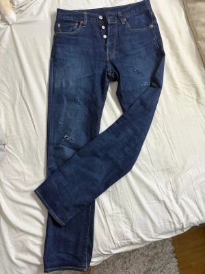 Levis 501 jeans  - Snygga mörkblå 501 jeans med raka ben och slitna detaljer på framsidan. Klassisk femficksmodell med knäppning fram och kontrastsömmar. Jeansen har en cool vintagekänsla och passar perfekt till en avslappnad stil.