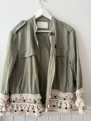 Grön overshirt med virkade detaljer - Stilren grön overshirt med virkade och fransade detaljer i beige längs nederkant och ärmslut. Jackan har två bröstfickor, knappar framtill och en avslappnad passform. Perfekt för dig som vill ha en unik touch på din outfit OBS skulle säga att den är mer än S än M!.