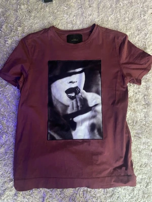 Vinröd t-shirt med tryck - Vinröd t-shirt med ett stort svartvitt tryck av ett ansikte på framsidan. T-shirten har rund hals och korta ärmar. Materialet känns mjukt och passar perfekt till en avslappnad stil. Litet hål men syns knappt vid användning