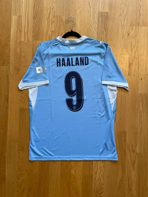 Haaland #9 - L - Snygg ljusblå Manchester City matchtröja från Puma med HAALAND och nummer 9 på ryggen. Tröjan har vita detaljer på ärmarna, klubbmärke och sponsortryck på bröstet samt rund hals. Perfekt för fotbollsfans som vill stötta sitt lag.