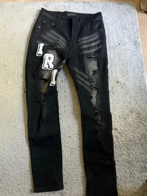Svarta distressed jeans med patches - Säljer ett par svarta jeans med slitningar och vita bokstavspatches på både fram- och baksida. Jeansen har skinny fit, fem fickor och coola detaljer som ger en streetwear vibe. Materialet är denim med stretch för extra komfort.
