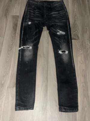 Svarta slitna jeans från Jack & Jones - Säljer ett par svarta jeans från Jack & Jones med slitningar och hål på båda benen. Jeansen har skinny fit och coola detaljer som blekta partier och färgstänk. Perfekta för dig som gillar en trendig och avslappnad stil.
