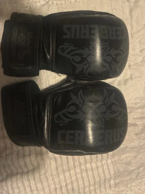 Svarta MMA-handskar från Cerberus - Säljer ett par svarta MMA-handskar från Cerberus med öppna fingrar och vaddering över knogarna. Handskarna har kardborreknäppning vid handleden och är tillverkade i slitstarkt syntetmaterial. Perfekta för kampsportsträning och sparring.