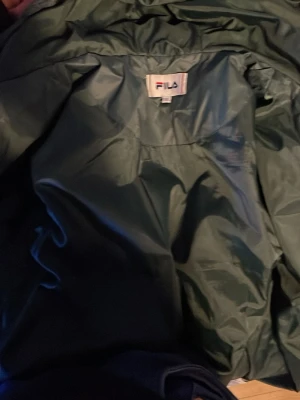 Mörkgrön pufferjacka från FILA - Säljer en mörkgrön pufferjacka från FILA med stor logga på bröstet. Jackan har glansig yta och är vadderad för extra värme. Perfekt för kalla dagar och har en klassisk pufferstil som är både trendig och bekväm.