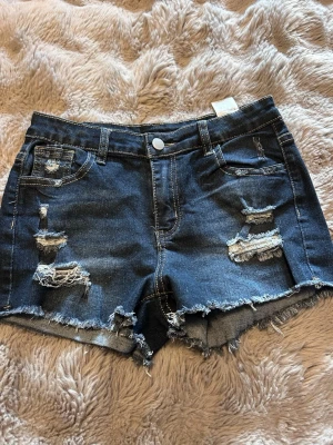 Mörkblå slitna jeansshorts - Säljer ett par mörkblå jeansshorts med slitna detaljer och råa kanter. Shortsen har klassisk femficksdesign, bälteshällor och knappstängning framtill. Perfekta för varma dagar och ger en avslappnad vibe.