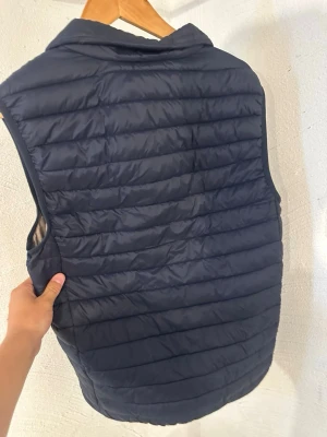 Mörkblå quiltad dunväst - Snygg mörkblå dunväst med quiltad design och diskreta horisontella sömmar. Västen har en enkel, stilren look och en liten ögla med orange och vit detalj i nacken. Perfekt för lager-på-lager och passar till många outfits.