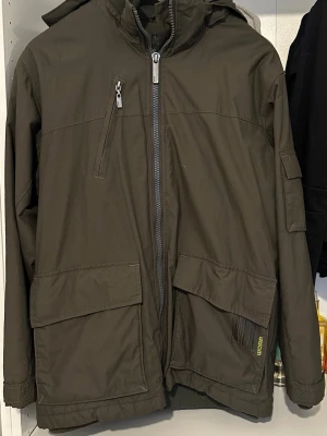 Grön jacka  - Snygg grön jacka med dragkedja, stor huva och flera praktiska fickor fram och på ärmen. Jackan har en diskret logga i grönt och är tillverkad i ett tåligt, vattenavvisande material. Perfekt för kyliga dagar och har en modern, avslappnad passform.