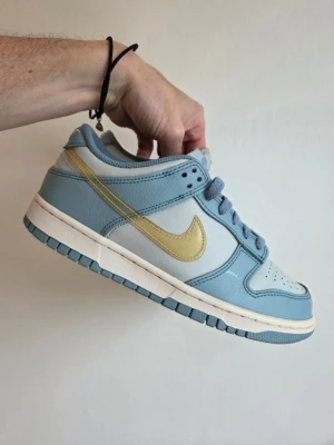 Nike Dunk Low blå/gul sneakers - Snygga Nike Dunk Low sneakers i ljusblått och vitt med en gul swoosh på sidan. Skorna har klassisk låg siluett, matchande blå snörning och vit mellansula. Perfekta för dig som vill sticka ut med stilrena färger och ikonisk design.