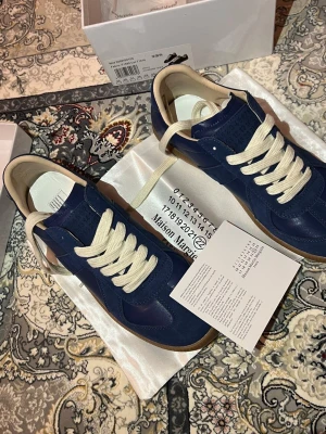 Blå Maison Margiela sneakers - Snygga blå sneakers från Maison Margiela med ovandel i både mocka och läder. Skorna har vita, breda snören och en klassisk gummisula i brunt. Låg modell med stilren design och diskreta detaljer. Perfekta för dig som vill ha något unikt och trendigt.