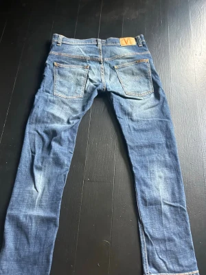 Nudie jeans blå - Nudie Jeans ”Thin Finn” i färgen Indigo Dreams. Modellen har en slim fit-passform med smala ben och normal midjehöjd. Mörk indigofärg med snygga, naturliga fades på låren och bakom knäna.  Storlek: W32 L30 Material: 99 % ekologisk bomull och 1 % elastan. Detaljer: Dragkedjegylf och femficksmodell.  Skicket är mycket bra, utan hål eller fläckar. Jeansens använda look syns genom tvätten på ett stilrent sätt.  Skickas inom 2 dagar. Hör gärna av dig om du har några frågor!