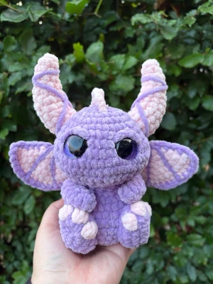 Lila virkad drake💜 - Supergulligt gosedjur i form av en drake, virkad i lila och ljusrosa plyschgarn. Draken har stora glittriga svarta ögon, stora öron och vingar samt små tassar. Perfekt som dekoration eller present till någon som gillar fantasy och söta saker. Rekommenderar ej till småbarn då säkerhetsögonen kan utgöra en kvävningsrisk!
