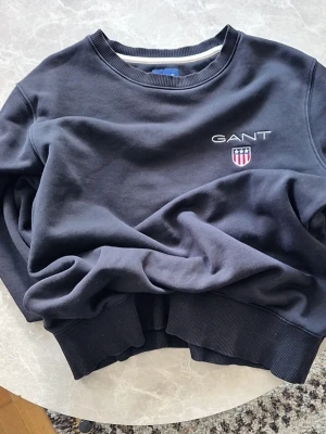 Svart tröja från GANT storlek M - Klassisk svart sweatshirt från GANT med broderad logga och sköld på bröstet. Tröjan har rund halsringning och ribbade muddar vid ärmslut och nederkant. Perfekt för en avslappnad och stilren look.