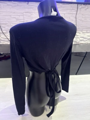 Svart croppad topp med knyt i ryggen - Snygg svart croppad topp med långa ärmar och knyt i ryggen. Toppen har en enkel, stilren design och är perfekt för att skapa en trendig look. Tillverkad i mjukt material som sitter skönt mot kroppen.
