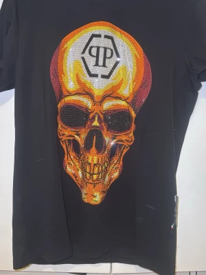 Philip plein T-shirt  - Cool svart t-shirt med ett stort, färgstarkt dödskalletryck i orange, gult och rött på framsidan. Trycket har en glansig effekt och ett grafiskt märke upptill. Perfekt för dig som gillar statement-plagg och streetwear.