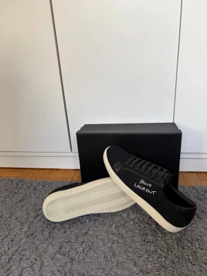 Svarta Saint Laurent sneakers - Snygga svarta sneakers från Saint Laurent med vit sula och handskriven logga på sidan. Skorna har klassisk låg profil, svarta skosnören och är tillverkade i canvas med detaljer i läder. Perfekta för dig som gillar stilrena och exklusiva sneakers.Skorna endast använda 1 gång. Skickar inom 24 timmar. Hör av er vid frågor!👋