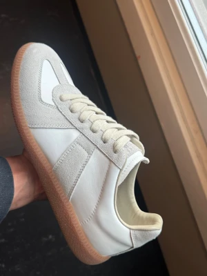 Maison Margiela Vita size 42  - Säljer ett par stilrena sneakers i vitt med beige mockadetaljer och klassisk gummisula. Skorna har låga skaft, vita snören och en retroinspirerad design som passar perfekt till jeans eller cargo. Materialet är en mix av slätt läder och mocka för en schysst kontrast.