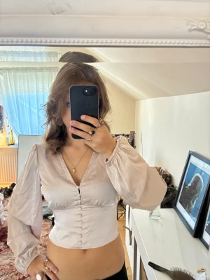 Beige satinblus med knappar - Snygg beige satinblus med djup v-ringning och dekorativa knappar framtill. Blusen har långärmade puffärmar med resår vid ärmslut och en figurnära passform som framhäver midjan. Perfekt för dig som gillar en trendig och feminin look.