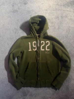 Vintage hollister hoddie  - Säljer en olivgrön hoodie med dragkedja och stor broderad text '1922' samt 'HCo' på bröstet. Hoodien har två fickor framtill, justerbar huva med snören och mjukt innerfoder. Perfekt för chill dagar eller när du vill ha en avslappnad stil.