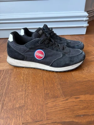 Svarta Colmar sneakers i mocka - Snygga svarta sneakers från Colmar med ovandel i mocka och textil. Skorna har vita detaljer vid hälen, svart snörning och en vit sula. På sidan sitter Colmars klassiska runda logga i rött, vitt och blått. Perfekta för en sportig och trendig look. Storlek 43. Bra skick