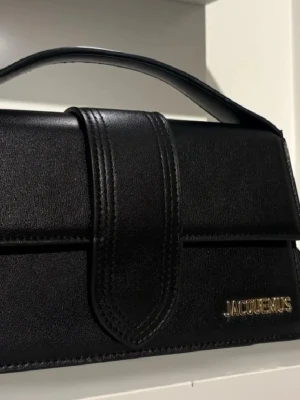 Svart Jacquemus handväska i läder - Snygg svart handväska från Jacquemus i slätt läder med guldfärgad logga framtill. Väskan har ett stilrent, rektangulärt format och ett kort handtag upptill. Perfekt accessoar för att lyfta vilken outfit som helst.