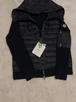 Svart cardiganjacka från Moncler - Snygg svart cardiganjacka från Moncler med quiltad front och huva. Jackan har stickade ärmar, dragkedja framtill och en liten ficka med dragkedja på ena ärmen. Perfekt för lager-på-lager och streetstyle.
