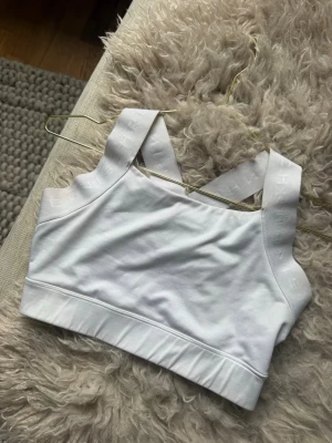 Vit sport-bh med breda axelband - Säljer en vit sport-bh med breda, korsade axelband och elastisk kant nedtill. Bh:n är tillverkad i ett stretchigt material och har diskret mönster på banden. Perfekt för träning eller yoga.