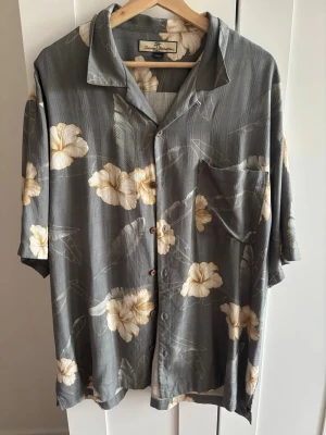 Blommig skjorta - Snygg skjorta av 100% silke med beige blommönster och korta ärmar. Skjortan har en avslappnad passform, klassisk krage och knäppning framtill. 