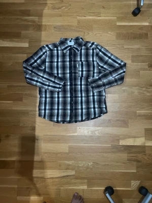 Svartvit rutig skjorta med knappar - Säljer en svartvit rutig skjorta med långa ärmar och klassisk krage. Skjortan har knappar framtill och en bröstficka. Perfekt för dig som gillar en avslappnad men ändå stilren look.