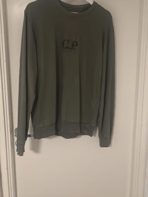 Olivgrön sweatshirt från C.P. Company - Snygg olivgrön sweatshirt från C.P. Company med diskret logga på bröstet. Tröjan har rund halsringning, långa ärmar och ribbade muddar vid ärmslut och nederkant. Perfekt för en avslappnad och stilren look.