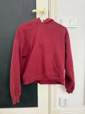 Mörkröd hoodie med magficka - Säljer en mörkröd hoodie med klassisk huva och stor magficka framtill. Hoodien har ribbade muddar vid ärmslut och nederkant. Perfekt för dig som gillar en enkel och stilren look. Materialet känns mjukt och bekvämt.