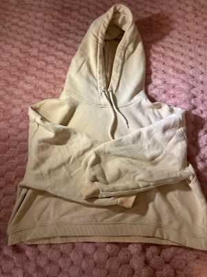 Beige hoodie med huva från 157 - Mysig beige hoodie från 157 med stor huva och dragsko. Insidan är fodrad med mjukt fleece-tyg för extra komfort. Klassisk passform med ribbade muddar vid ärmslut och nederkant. Perfekt för chill dagar eller när du vill ha något bekvämt.