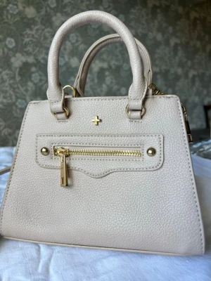 Beige handväska från PETA + JAIN - Stilren beige handväska från PETA + JAIN i strukturerat konstläder. Väskan har två handtag, guldfärgade metalldetaljer och en liten ficka med dragkedja framtill. Axelrem finnes för crossbody.  Perfekt för dig som gillar minimalistisk och trendig design. Ca 20x20 cm