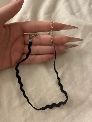 Svart vågig chokerhalsband - Säljer ett svart chokerhalsband med vågig design. Halsbandet har justerbar kedja i silverfärg och stängs med ett klassiskt spänne. Perfekt accessoar för att ge din outfit en edgy touch.