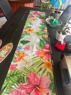 Färgglad tropisk dekoration  - Säljer en lång bordsduk med tropiskt blommönster i starka färger som rosa, gul, orange och grön. Perfekt för sommarfest eller temakväll. Materialet är plast och duken har ett livligt tryck av hibiskusblommor och palmblad.